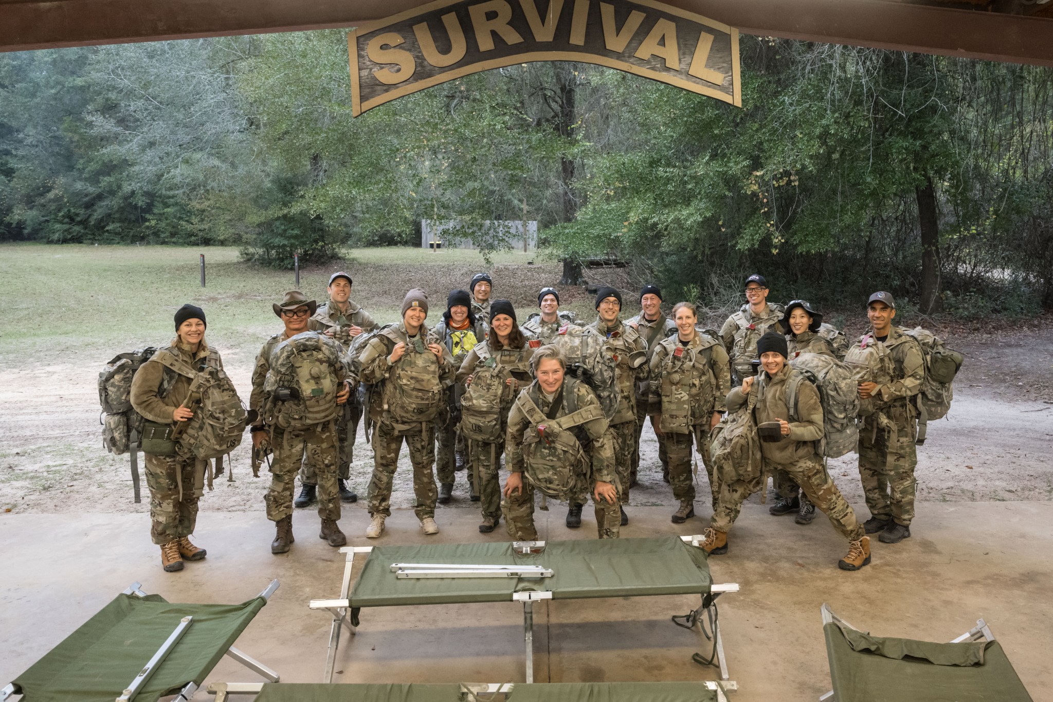 A group of people in camouflage uniforms and large backpacks stand together beneath a wooden “SURVIVAL” sign in a wooded outdoor training area, with green cots and a concrete shelter in the foreground.