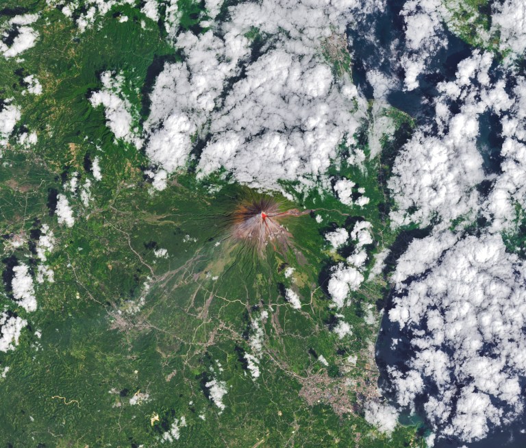 The upper slopes of Mayon volcano appear brown, with several narrow channels radiating from the crater. A red infrared heat signature appears near the summit, with red streaks extending east and southeast. The lower slopes are green and forested. Farmland and towns are visible in the lower part of the image.