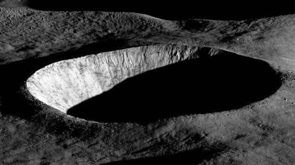Oblique view of a large, circular lunar crater with steep, rugged walls. Sunlight illuminates one side of the crater rim and inner wall, while the crater floor and much of the interior remain in deep shadow. The surrounding terrain appears gray and heavily textured with smaller craters and rocky ridges. The black sky of space fills the background.
