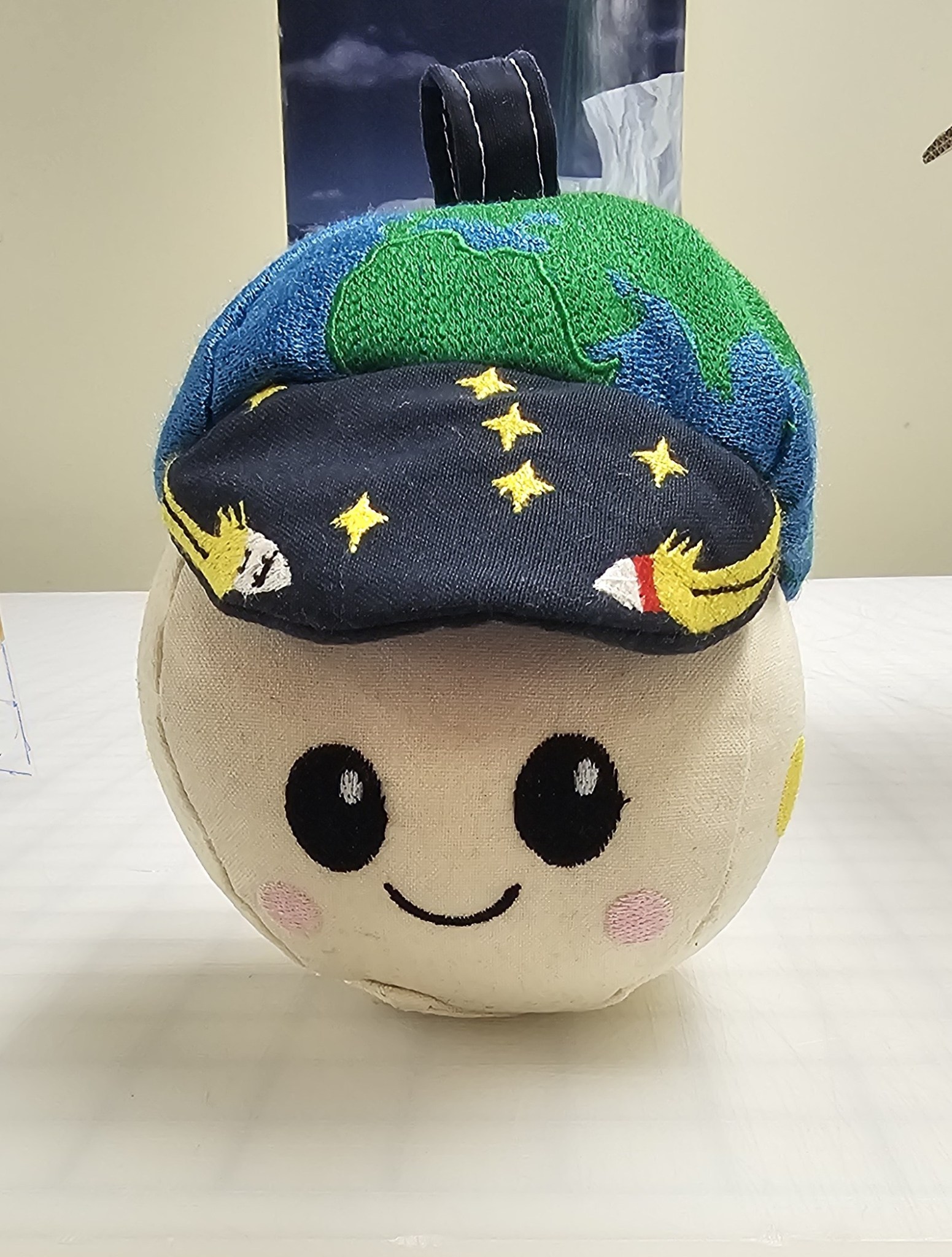 Image shows a white round plushy with a smiley face that has been sewn together with a hat designed in colors reflecting the Earth and a hat bill showing the galaxy with stars and rockets sewn on. Credit: NASA