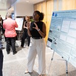 A person stands beside a large research poster on a tripod display, gesturing with both hands while speaking to another person who is standing nearby. The poster includes charts, graphs, and text under a NASA logo. Several other people are visible in the background viewing different posters in a brightly lit indoor hallway or event space.