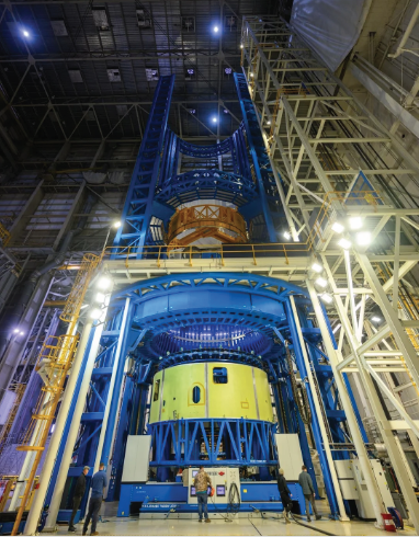 Interior of a tall industrial assembly building showing a large yellow‑green cylindrical aerospace structure held within a multi‑story blue steel support and processing tower. Surrounding the assembly are white and yellow access platforms, scaffolding, and bright overhead lighting. A few workers stand near the base, emphasizing the enormous scale of the structure