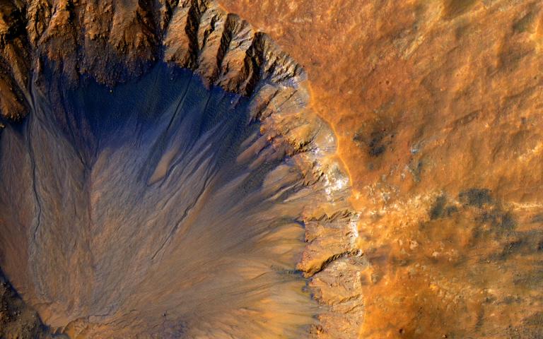 An impact crater with a sharp rim seen from above. Most of the landscape is orange, while the steep inner slope on the north side of the crater is a deep blue-black.