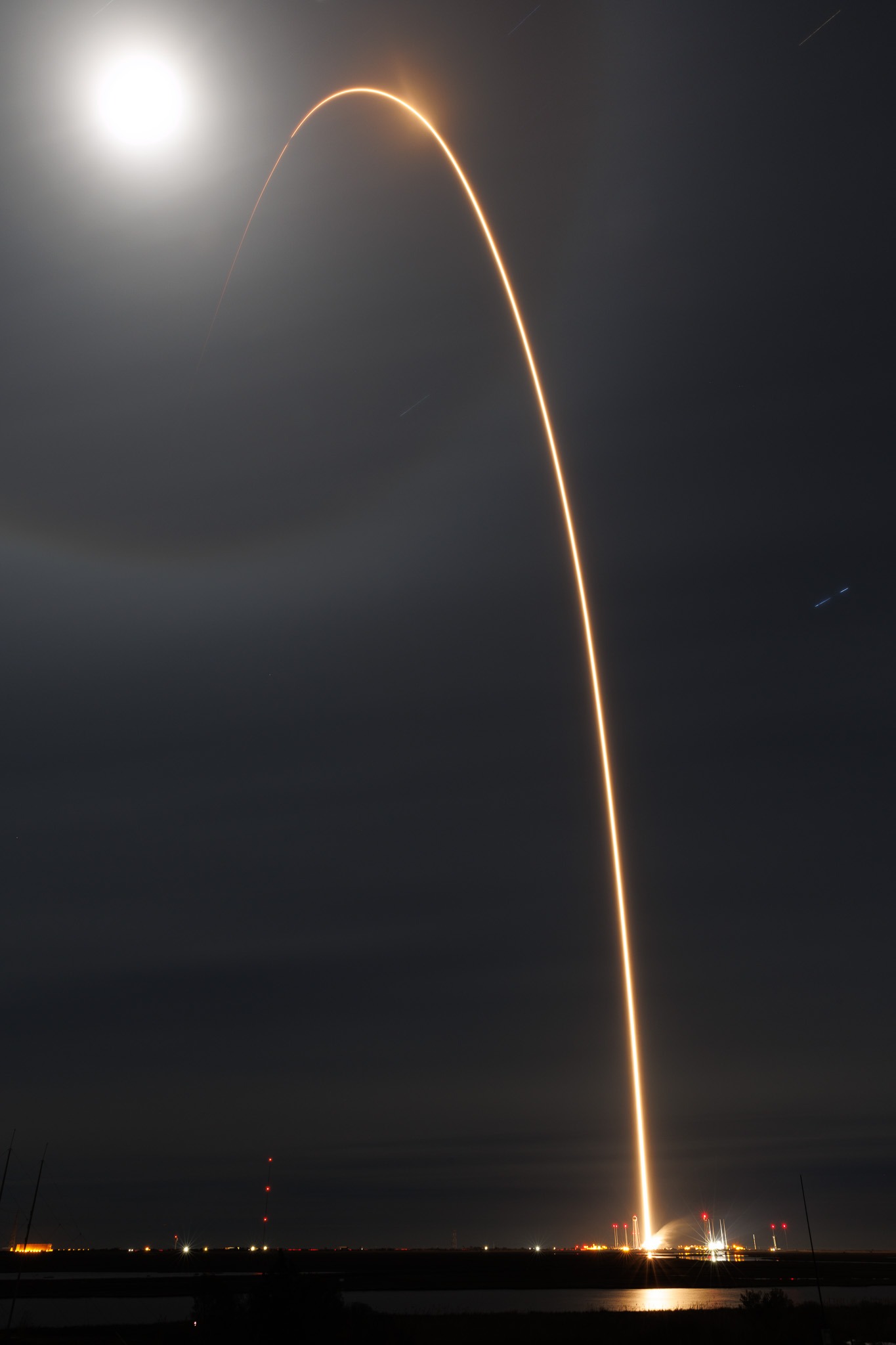 A long exposure photograph of a HASTE launch, which is a white streak against a black sky with a blurry white circular that is the Moon in the left corner.
