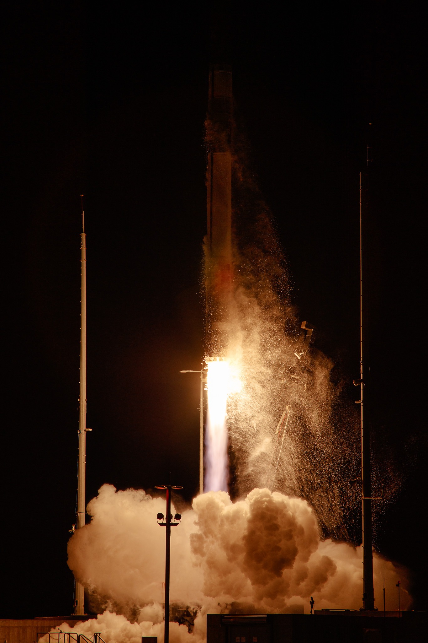 A Rocket Lab HASTE rocket lifts off at night from NASA's Wallops Island, with bright orange and white flames illuminating billowing clouds of smoke against a black sky. Launch structure towers are visible on either side of the rocket.