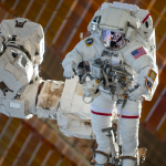 An astronaut in a white spacesuit is shown being maneuvered by the Canadarm2, which is attached to the space station, during a spacewalk.