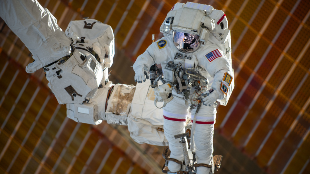 An astronaut in a white spacesuit is shown being maneuvered by the Canadarm2, which is attached to the space station, during a spacewalk.