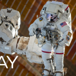 An astronaut in a white spacesuit is shown being maneuvered by the Canadarm2, which is attached to the space station, during a spacewalk.