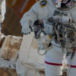 An astronaut in a white spacesuit is shown being maneuvered by the Canadarm2, which is attached to the space station, during a spacewalk.
