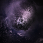 Lightening is shown flashing through wispy storm clouds off the coast of North Carolina while the crew was orbiting 260 miles (418 kilometers) above the Atlantic Ocean. The hue of the clouds is a mix of lighter and darker purples. The clouds appear to be funneling in towards a brighter mass of clouds in the center.