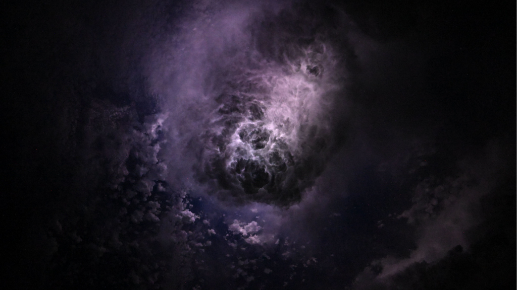 Lightening is shown flashing through wispy storm clouds off the coast of North Carolina while the crew was orbiting 260 miles (418 kilometers) above the Atlantic Ocean. The hue of the clouds is a mix of lighter and darker purples. The clouds appear to be funneling in towards a brighter mass of clouds in the center.
