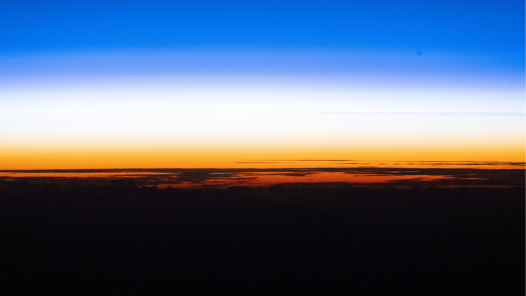 An orbital sunset silhouettes the cloud tops above the Pacific Ocean as the atmosphere along the horizon fades from orange to white, then to blue, in this photograph taken from the International Space Station while it orbited 262 miles above Earth.