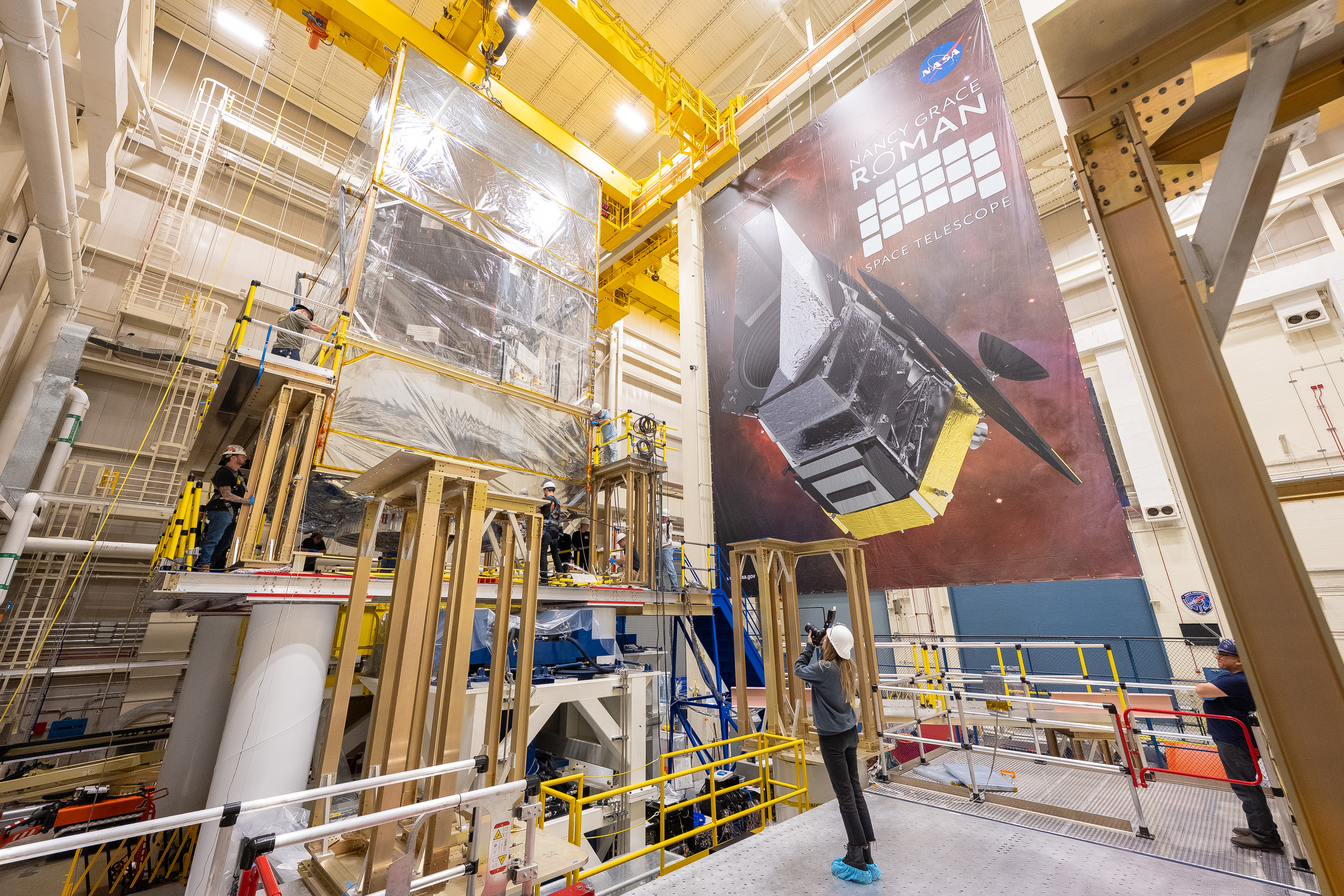 A woman in a white hard hat aims a camera up at a raised platform, onto which the Roman observatory is being lowered. Several people in hard hats surround the observatory to facilitate the process.