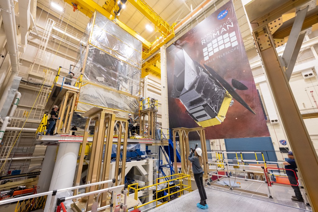 A woman in a white hard hat aims a camera up at a raised platform, onto which the Roman observatory is being lowered. Several people in hard hats surround the observatory to facilitate the process.
