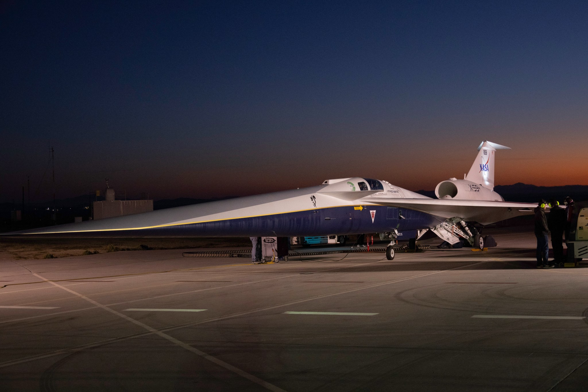 An aircraft resting on a section of runway as seen from the side. The X-59 has a long, thin nose that accounts for nearly a third of its length, along with sleek wings and an engine mounted above its body, just below its tail. The early morning sky is dark in the background with the sunrise just starting to emerge.