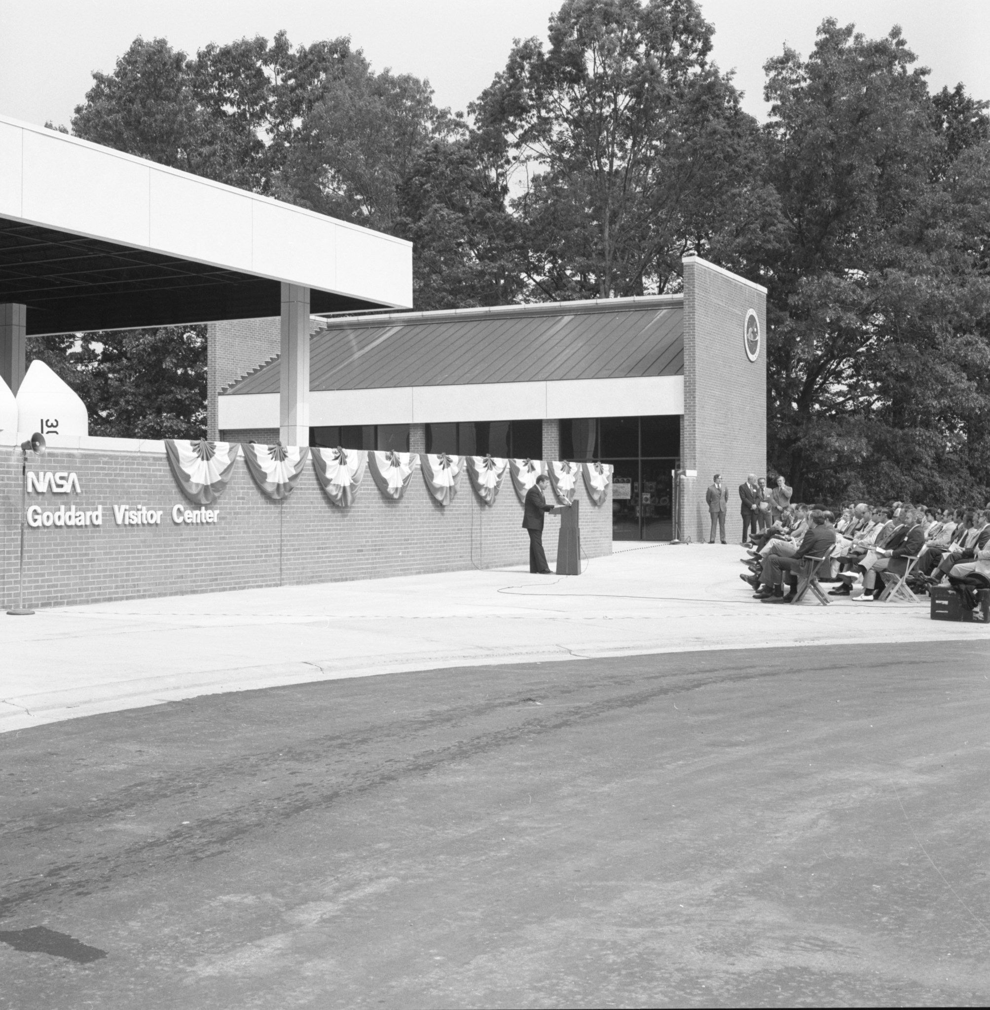 black and white photo of a man at a lectern addressing a small crowd in front of a brick building