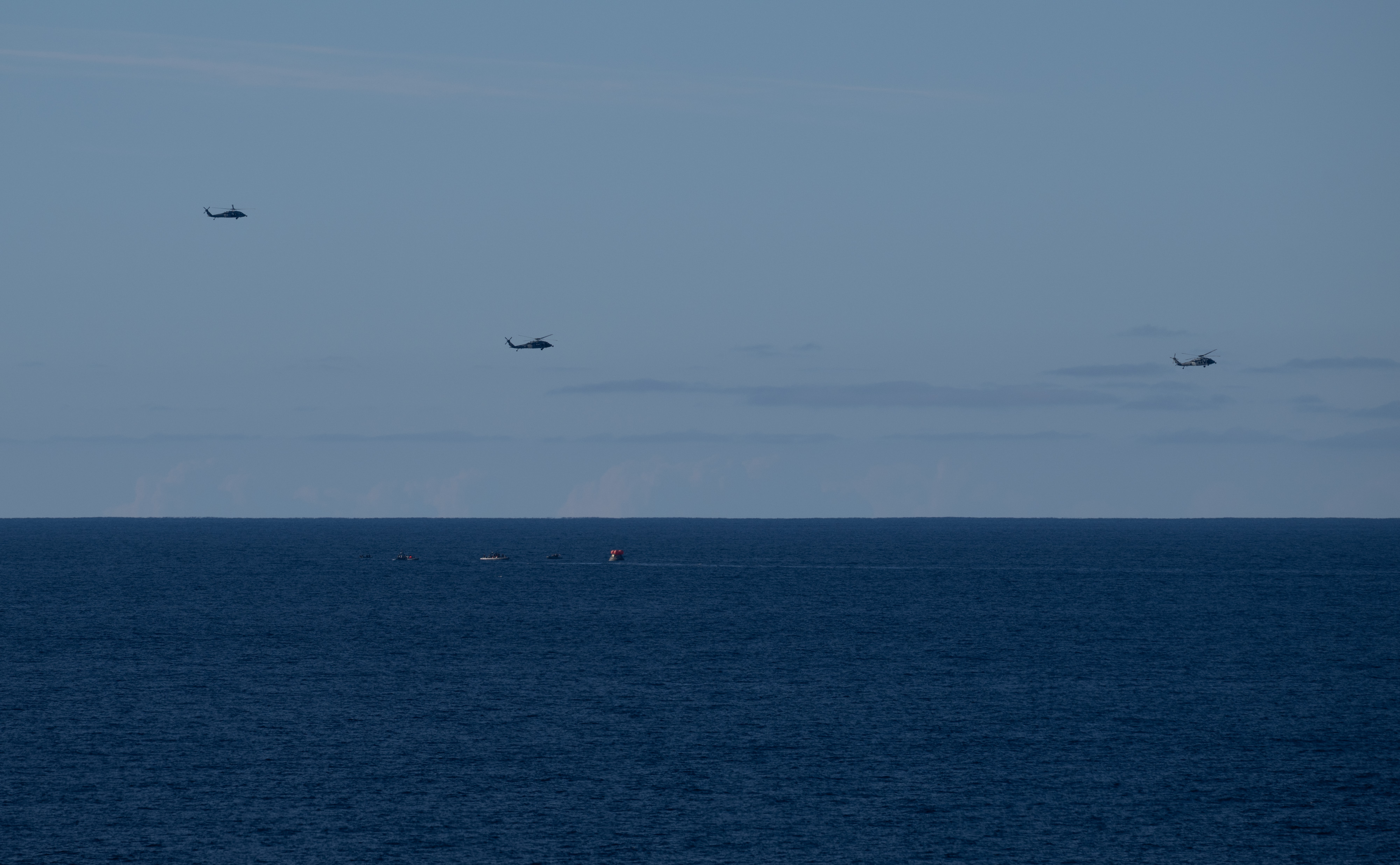 Three U.S. Navy MH-60 Seahawks from Helicopter Sea Combat Squadron (HSC) 23 flies overhead as small boats approach NASA’s Orion...