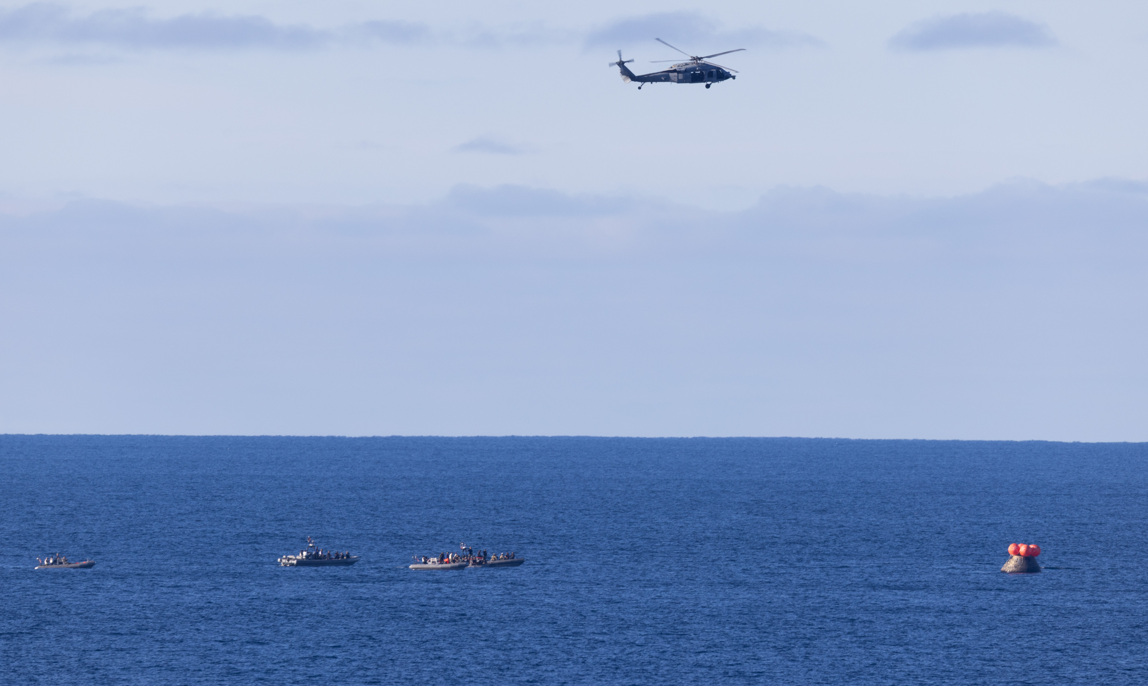 A U.S. Navy MH-60 Seahawks from Helicopter Sea Combat Squadron (HSC) 23 flies overhead as small boats approach NASA’s Orion...