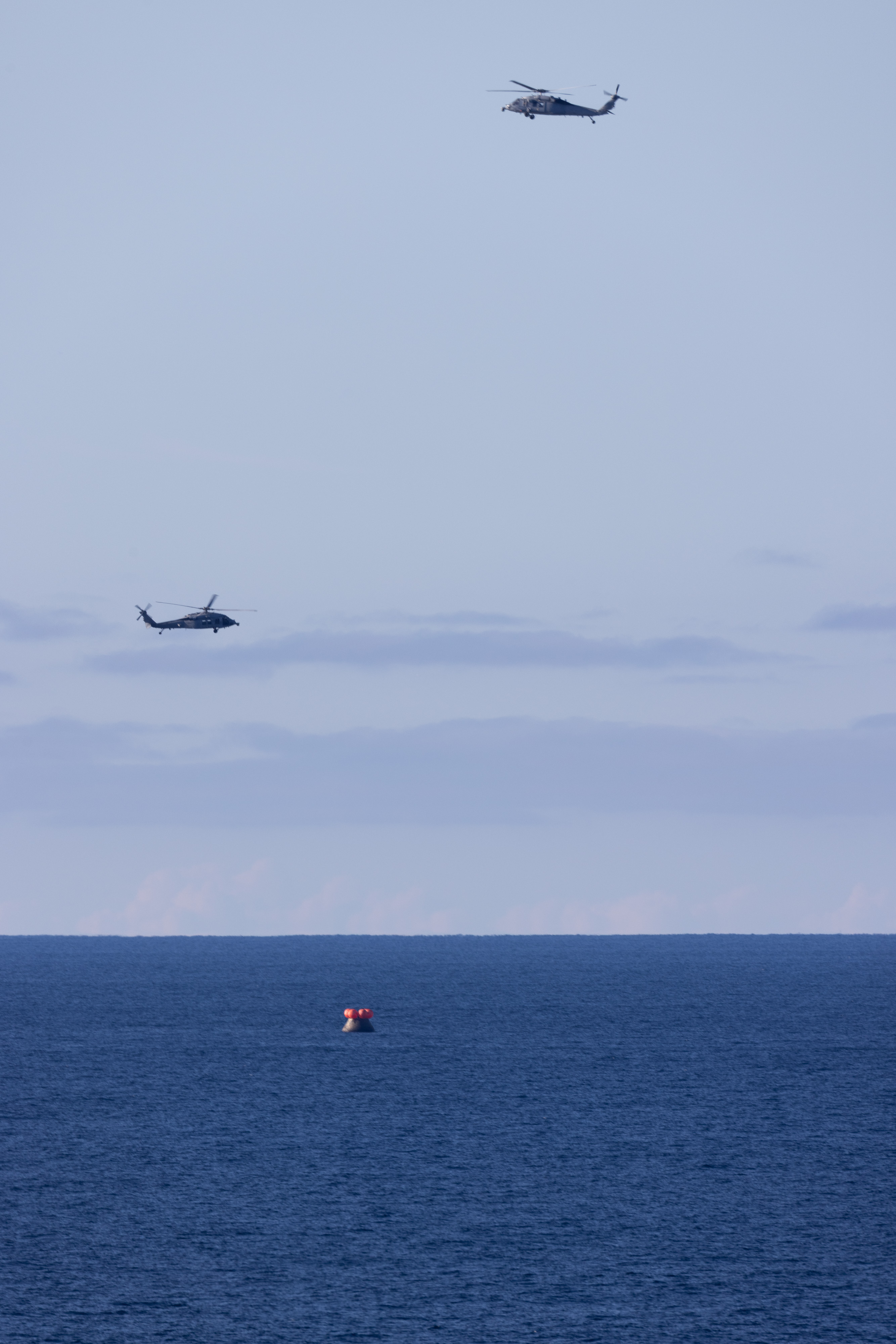 Two U.S. Navy MH-60 Seahawks from Helicopter Sea Combat Squadron (HSC) 23 flies overhead as small boats approach NASA’s Orion...