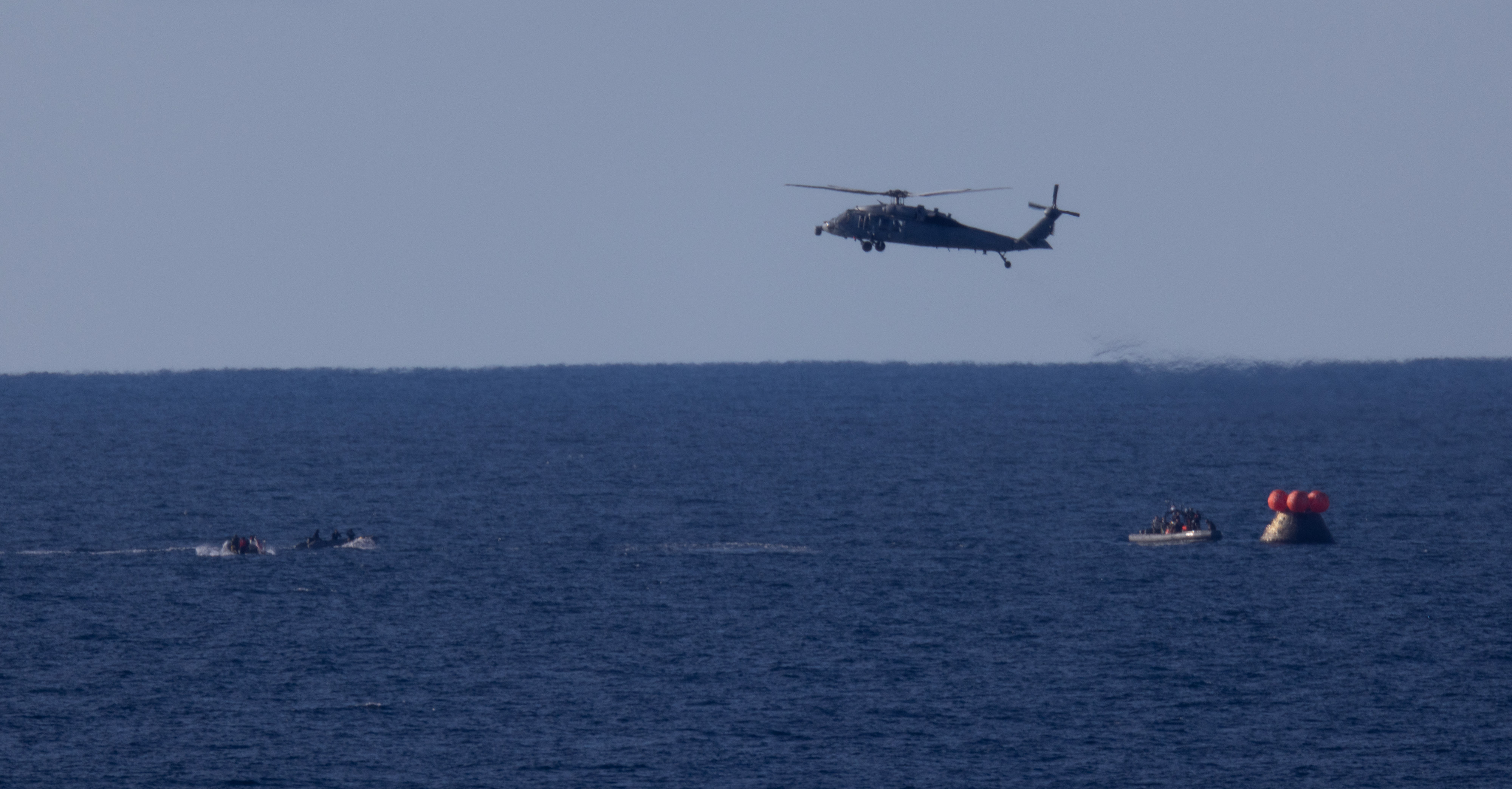 A U.S. Navy MH-60 Seahawks from Helicopter Sea Combat Squadron (HSC) 23 flies overhead as small boats approach NASA’s Orion...