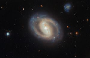 A face-on view of the barred spiral galaxy IC 486, showing a bright, elongated central bar and softly curving, ring-like spiral arms with subtle blue star-forming regions and dark dust lanes, set against a black background dotted with distant galaxies and a few foreground stars.