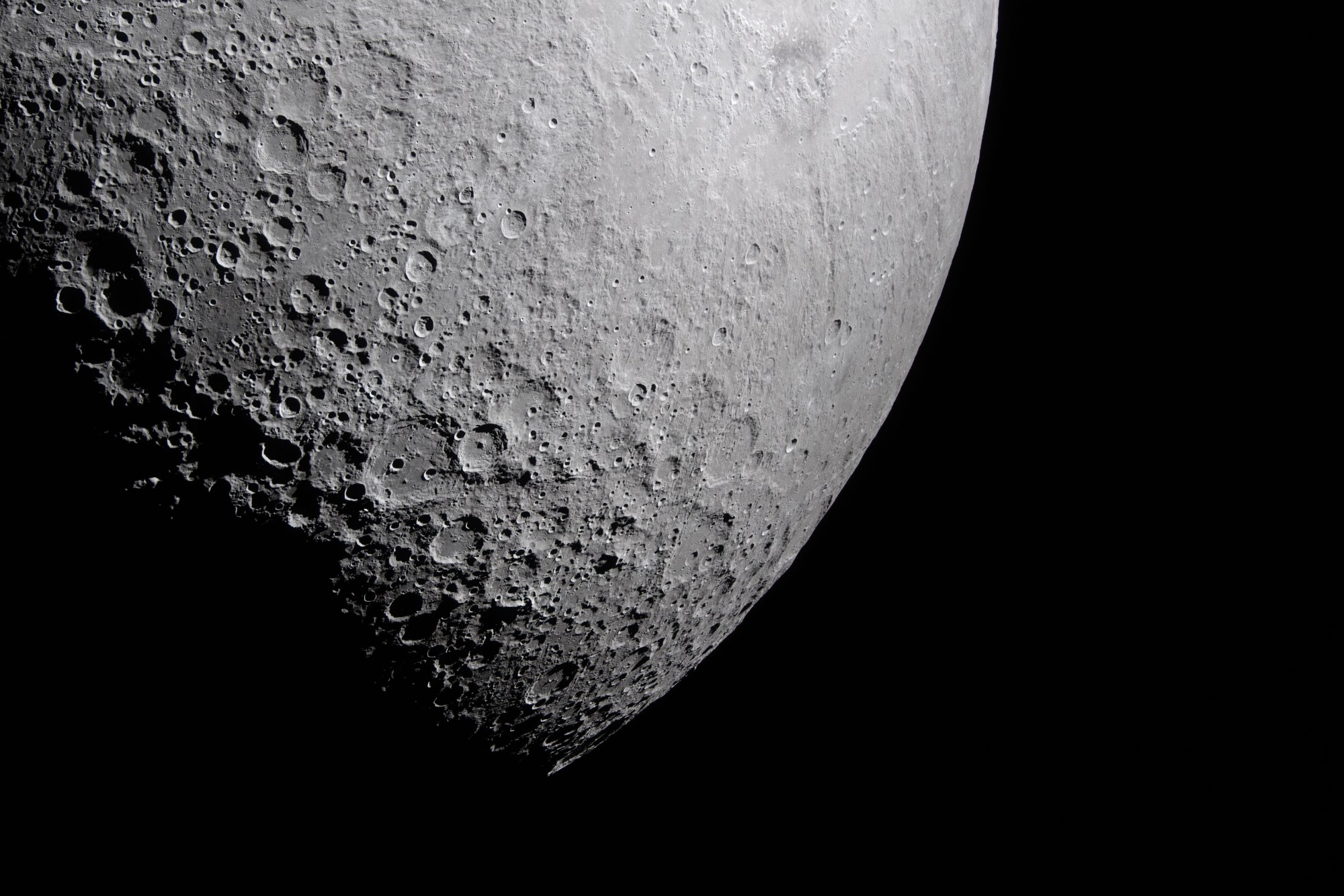 Part of the Moon stands out against the blackness of space. Its surface is marked with many craters of different sizes. The craters are mostly on the left and bottom. The upper right portion of the Moon's surface is much smoother.