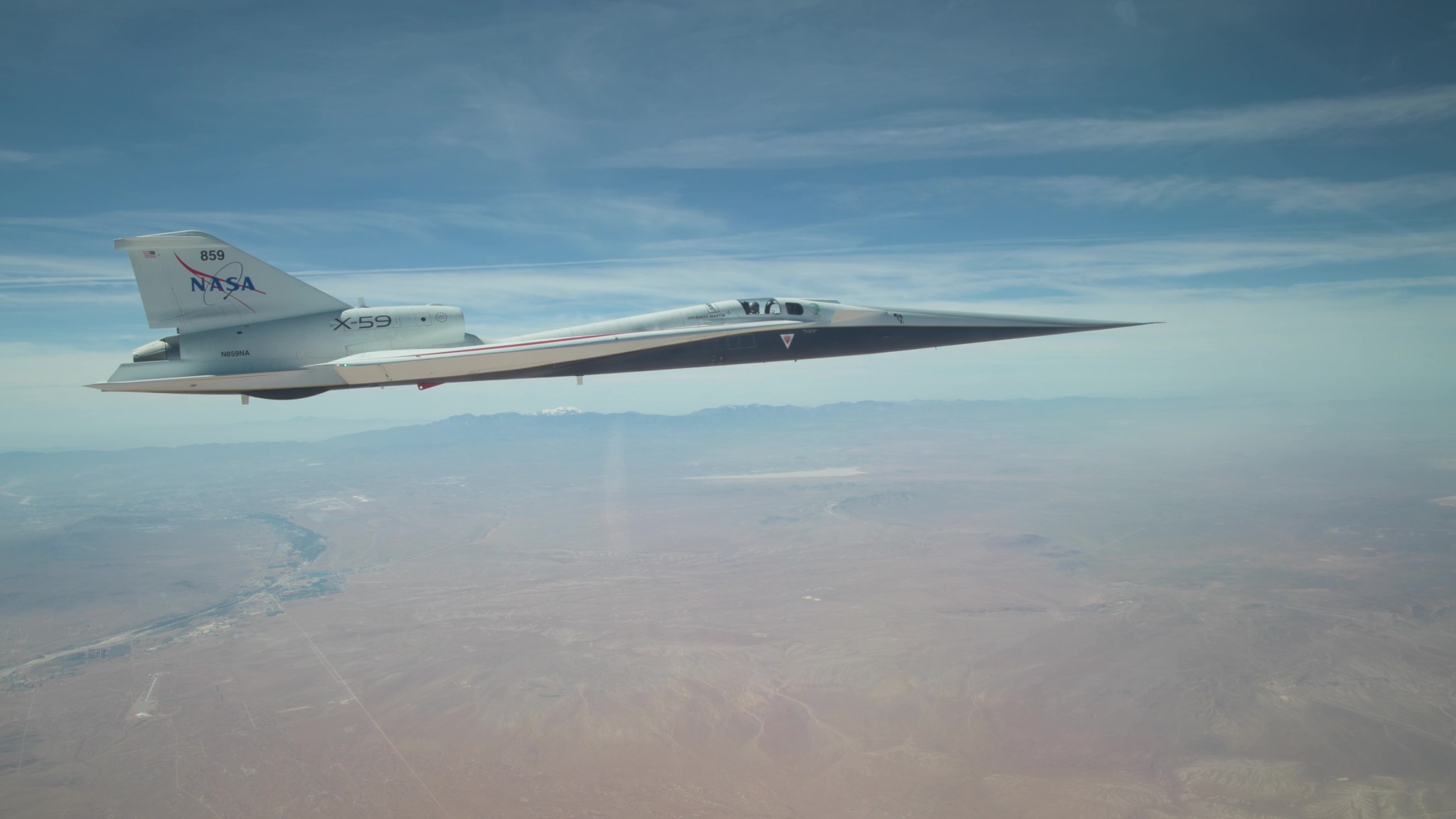 NASA’s X-59 quiet supersonic research aircraft flies above the Mojave Desert on a clear day. The aircraft is white with light gray, red, and blue accent colors. A NASA logo is visible on its tail, along with the number 859 above it. It appears to be flying level over the desert land-scape, and in the distance, you can see a mountain range on the horizon.