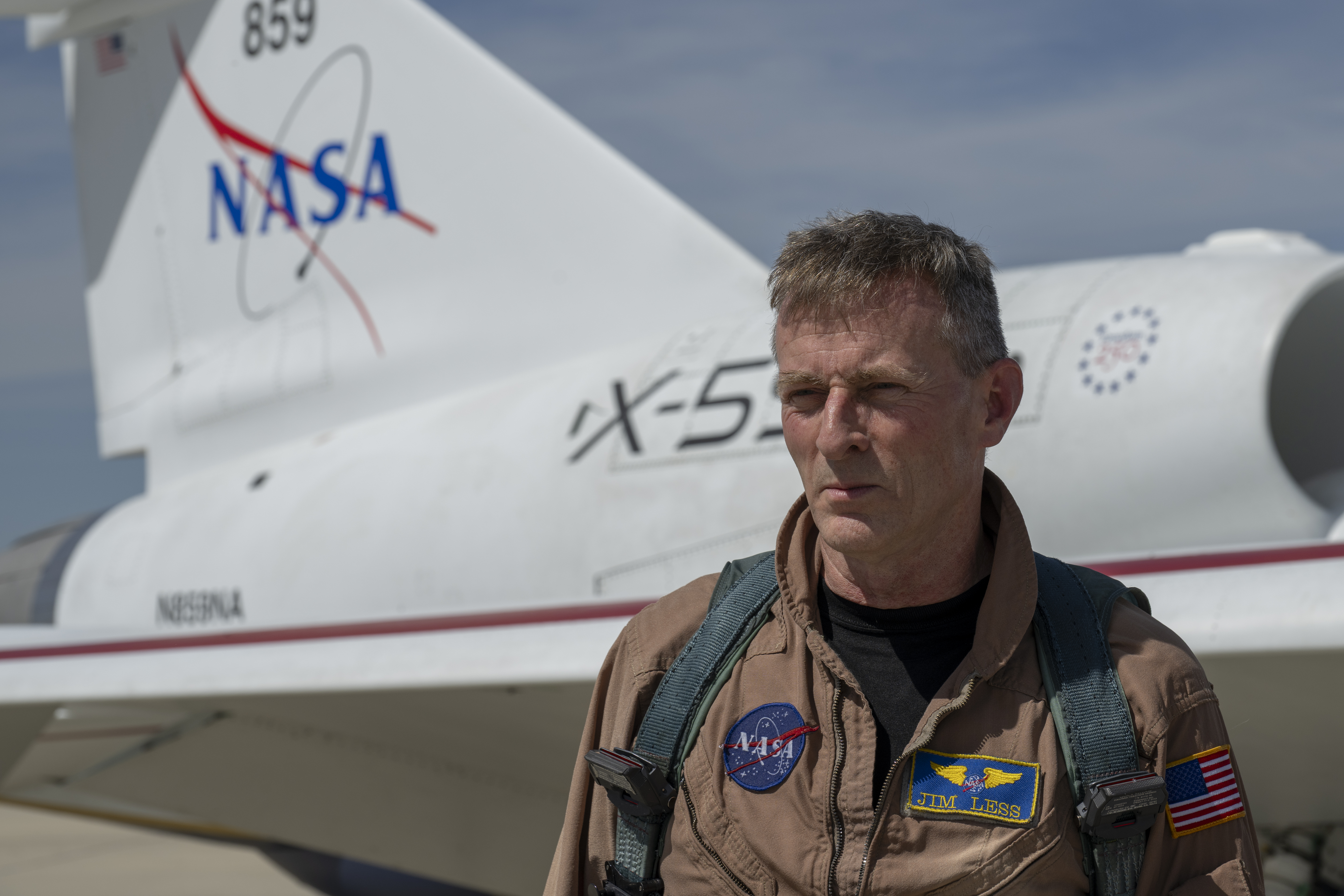 NASA Test Pilot Jim "Clue" Less wearing a tan flight suit with the X-59 behind him.