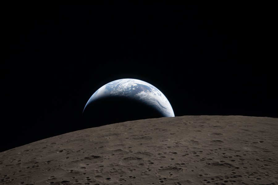 art002e009288 (April 6, 2026) – Earthset captured through the Orion spacecraft window at 6:41 p.m. EDT, April 6, 2026, during the Artemis II crew’s flyby of the Moon. A muted blue Earth with bright white clouds sets behind the cratered lunar surface. The dark portion of Earth is experiencing nighttime. On Earth’s day side, swirling clouds are visible over the Australia and Oceania region. In the foreground, Ohm crater has terraced edges and a flat floor interrupted by central peaks. Central peaks form in complex craters when the lunar surface, liquefied on impact, splashes upwards during the crater’s formation.