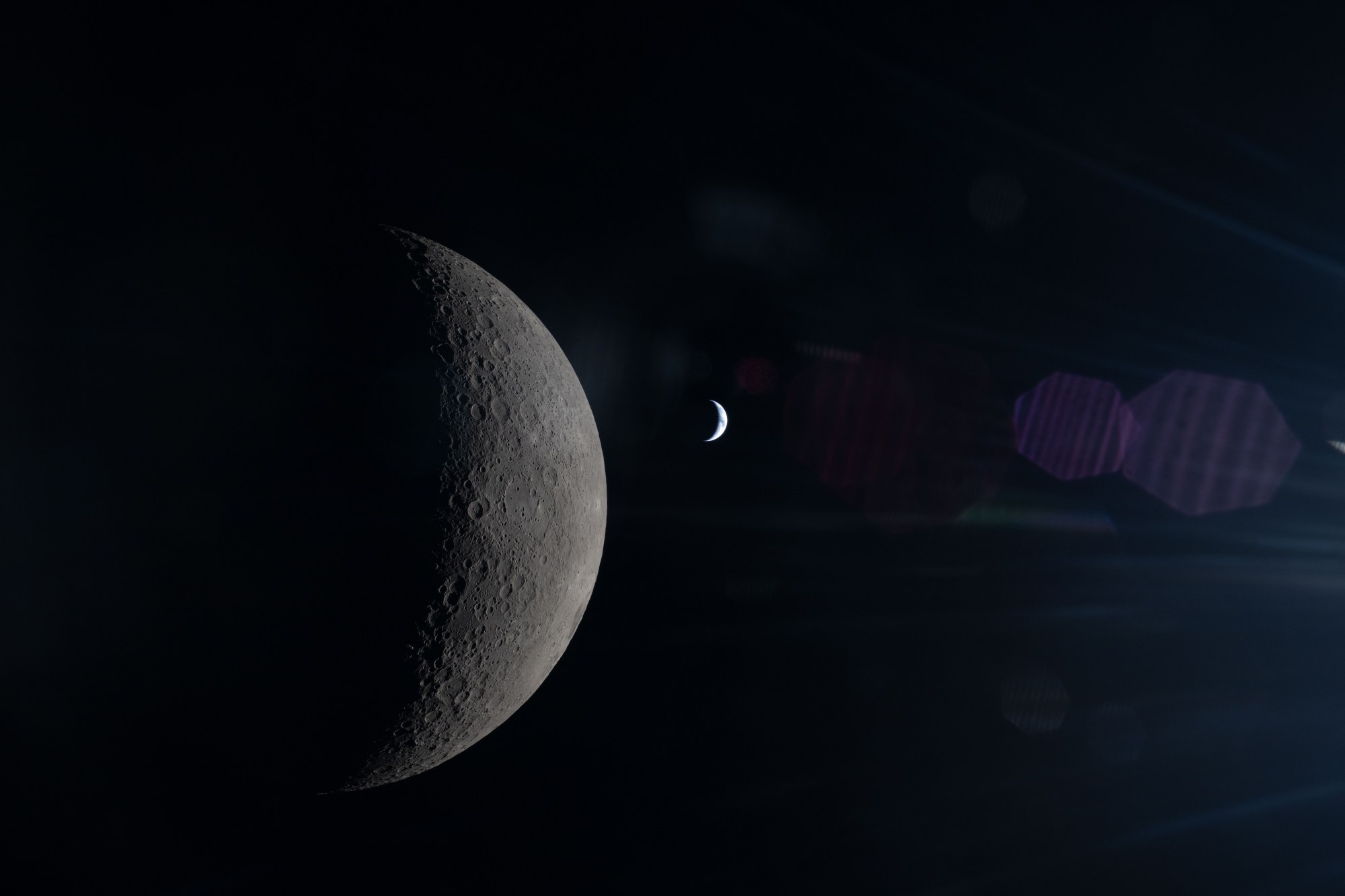 This image, taken from behind the Moon, shows its half-illuminated surface in sharp detail. To the right, a much smaller crescent Earth can be seen, with shades of blue and white just barely distinguishable. A faint reflection of the Orion spacecraft's window overlays the scene, especially in its right half.