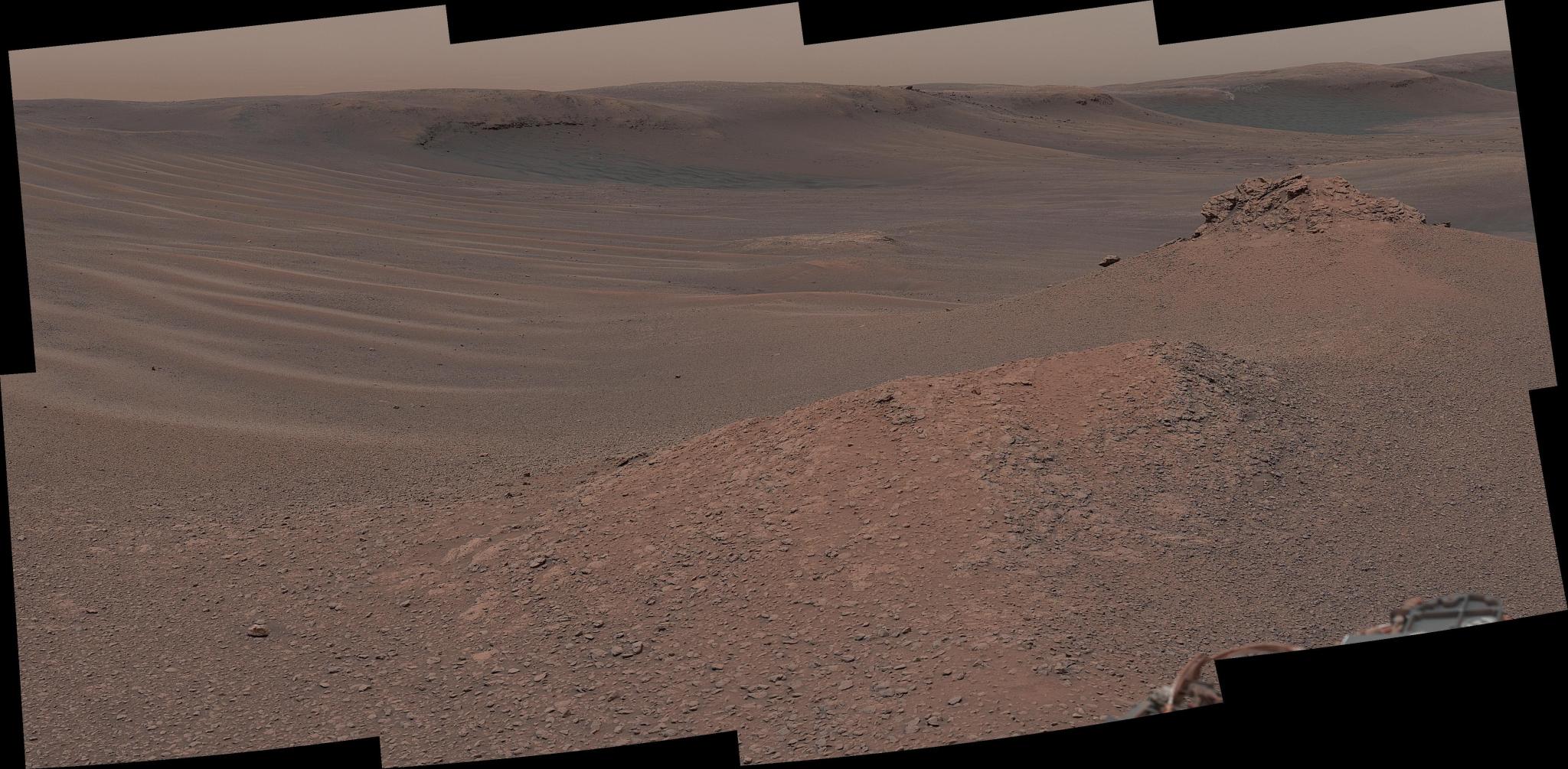 A vast, desolate Martian landscape features wind-swept sand ripples under a hazy salmon sky, with jagged rock formations in the foreground and rolling reddish-brown hills in the background.