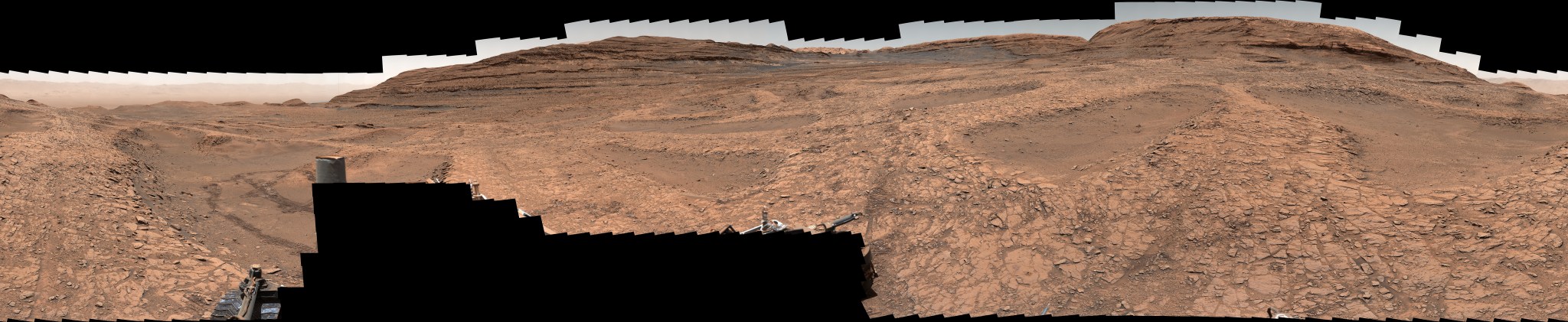 A series of shallow, sand-filled pits with low ridges spread across a tawny Martian landscape. Rover tracks stretch toward the horizon at left, and steep ridgetops loom in the background.