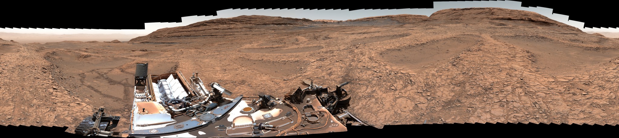 A rock-strewn, brownish-red Martian plain is bordered by a series of ridges and hilltops on the horizon, with rover tracks leading from them to the foreground on the right side of the image, where small portions of Perseverance rover are visible.