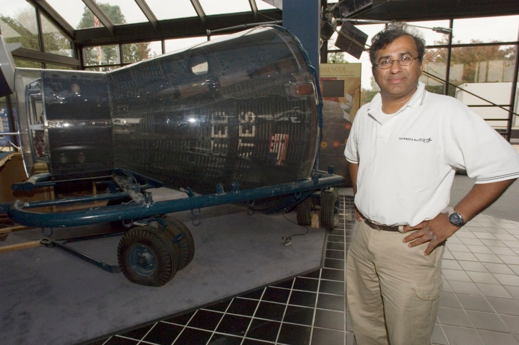 man in white polo stands next to a gray, plastic-encased Gemini space capsule