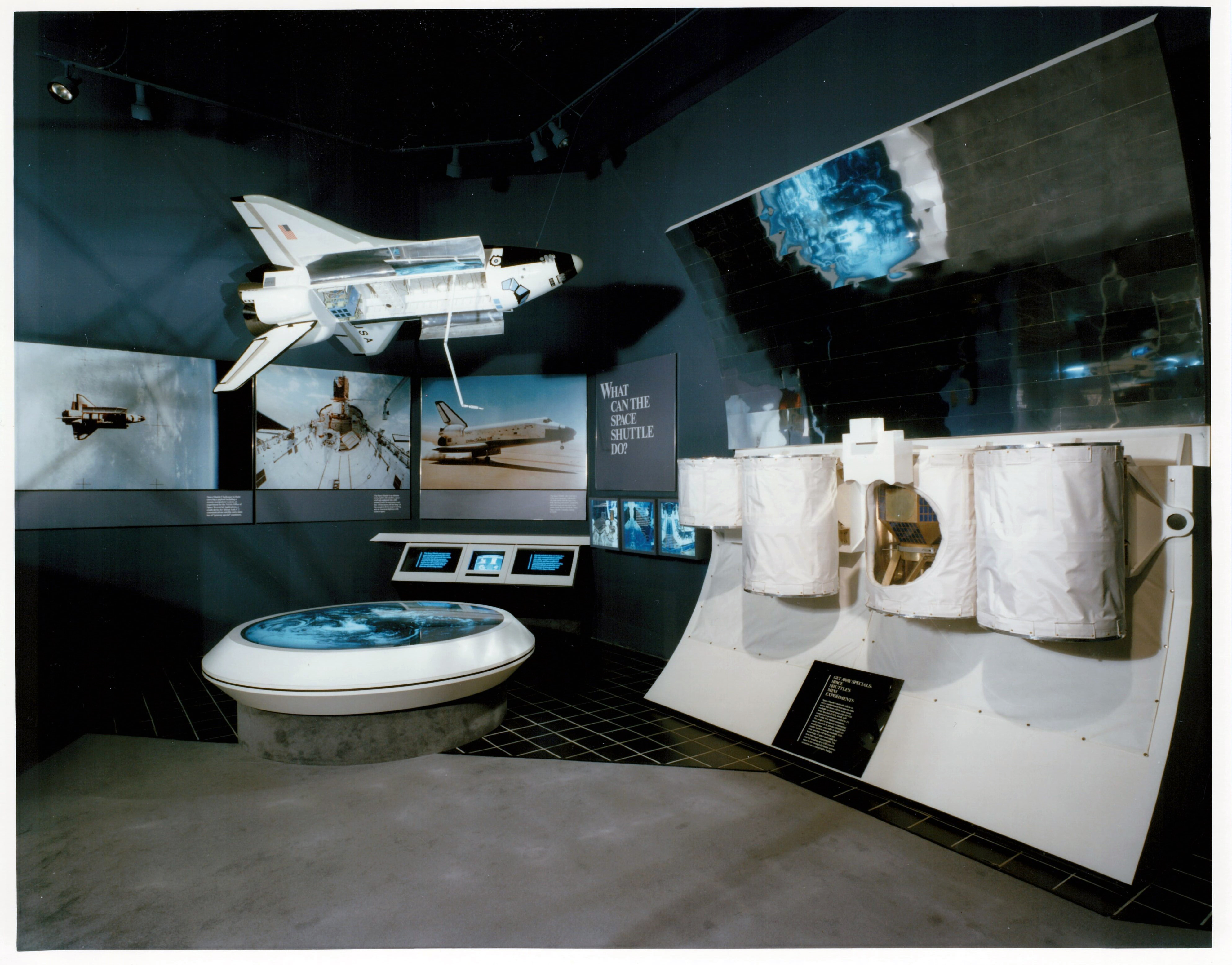 Small model of Space Shuttle hangs from the ceiling by an exhibit of small shuttle payloads shaped like white cylinders