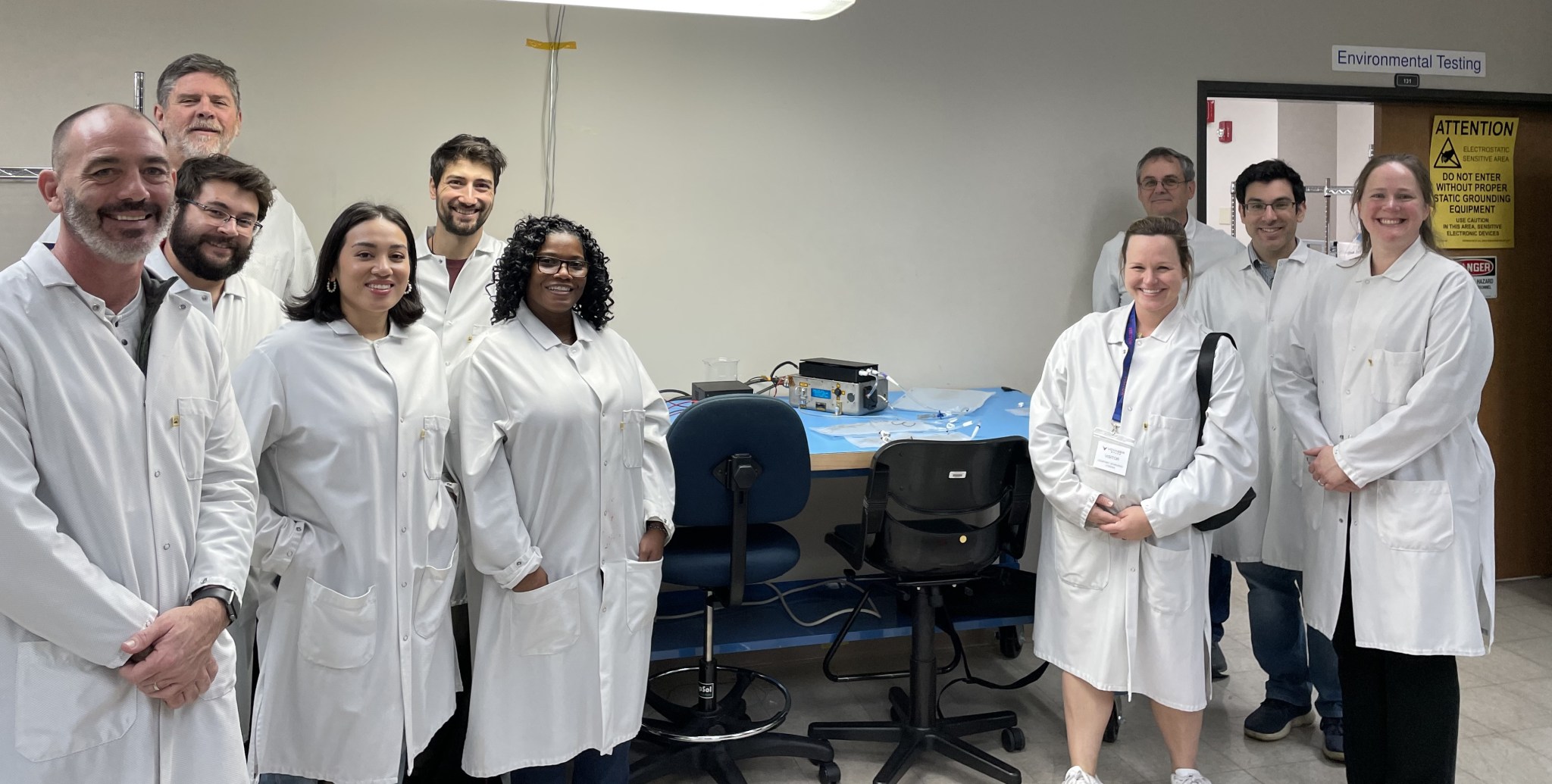A team of 10 people pose for a photo within a laboratory. The group includes four men and two women standing on the left side, all wearing lab coats. The IVGEN Mini system is on a table in the center of the photo, and to the right are four additional people standing – two women and two men.