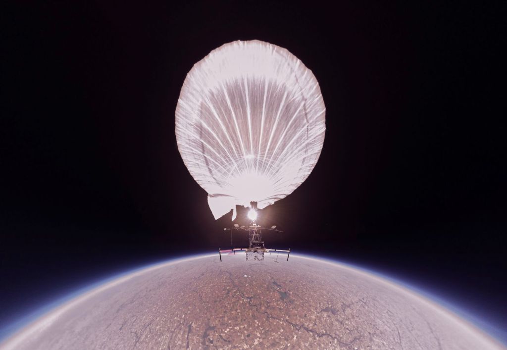 A photo of a high-altitude balloon payload suspended above Earth’s curved horizon, with a large white balloon illuminated against the dark upper atmosphere.