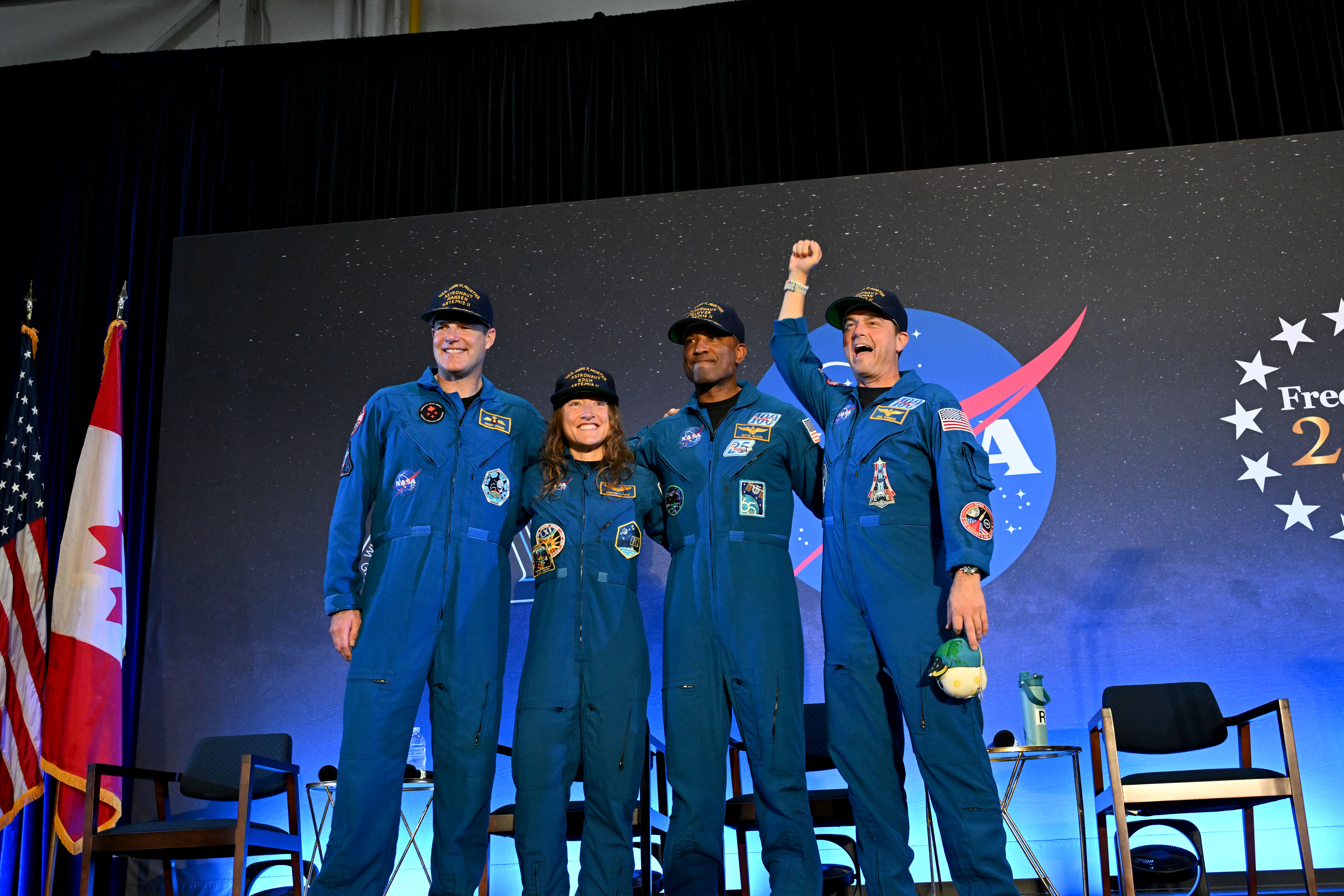 CSA (Canadian Space Agency) astronaut Jeremy Hansen and NASA astronauts Reid Wiseman, Victor Glover, and Christina Koch smile at the crowd during a news conference. They are all wearing blue jumpsuits with patches on the arms and chest areas. Wiseman raises his right fist in a victorious pose.