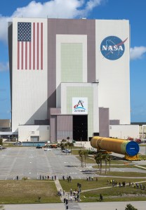 The Artemis III rocket core stage, a massive orange cylinder, moves into the tall Vehicle Assembly Building. A line of people watch from afar on a grassy lawn.