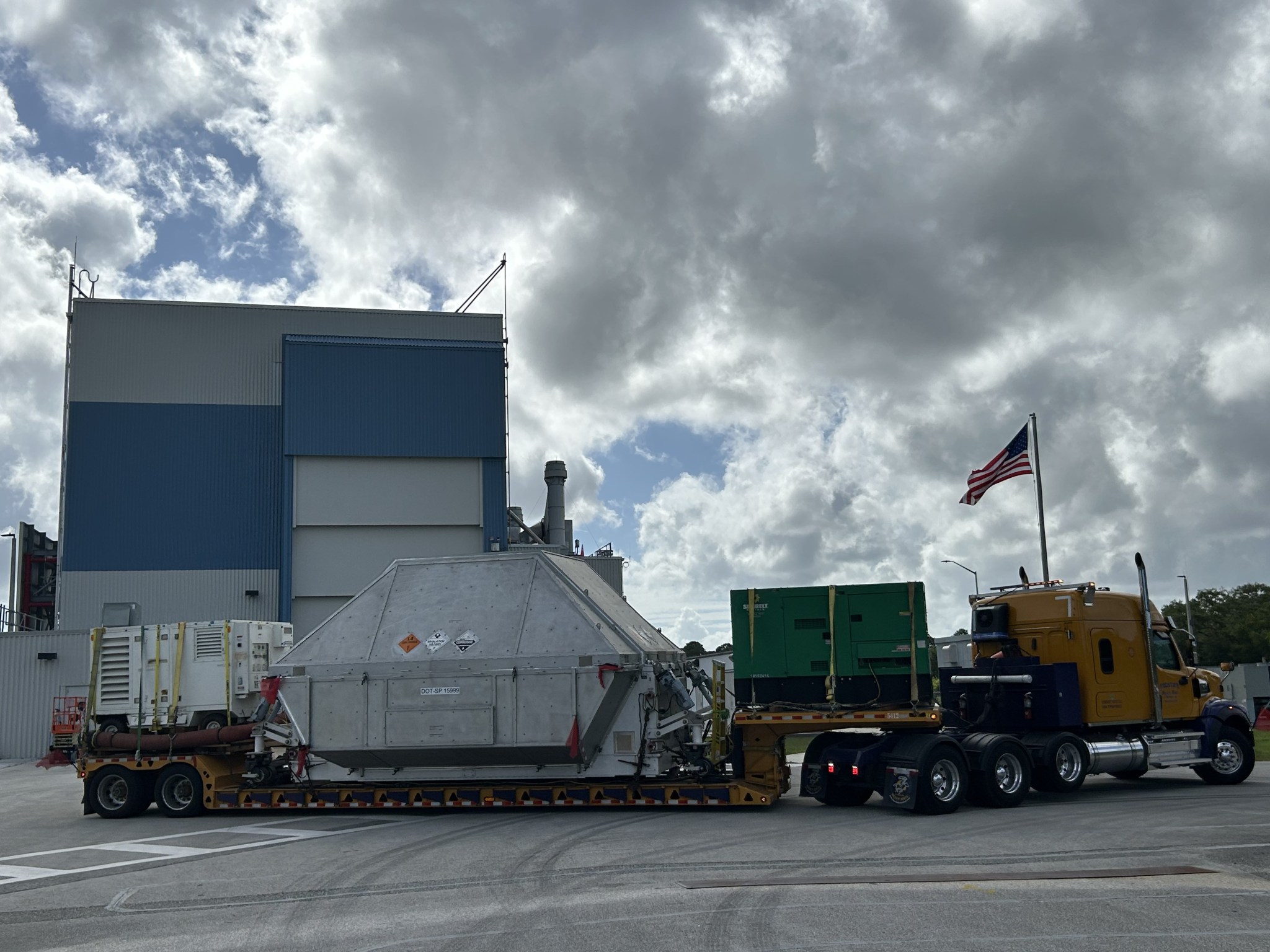 A large truck with a yellow cab hauls a wide, metal, container-like structure on a low trailer, with additional equipment secured behind it. An American flag flies from the truck as it sits on a paved area beside a tall industrial building. Thick, backlit clouds fill the sky above at NASA's Kennedy Space Center in Florida on Tuesday, April 28, 2026. Photo credit: NASA/Tiffany Fairley