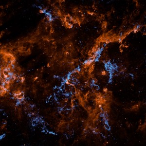 Wispy filaments of vibrant orange and electric blue cosmic dust and gas weave through a dark, star-studded expanse of outer space, creating a chaotic and intricate web of celestial matter.