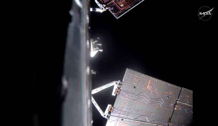 A view from an onboard camera showing parts of a spacecraft against the blackness of space. Large rectangular solar array panels with visible wiring patterns extend outward on articulated arms.