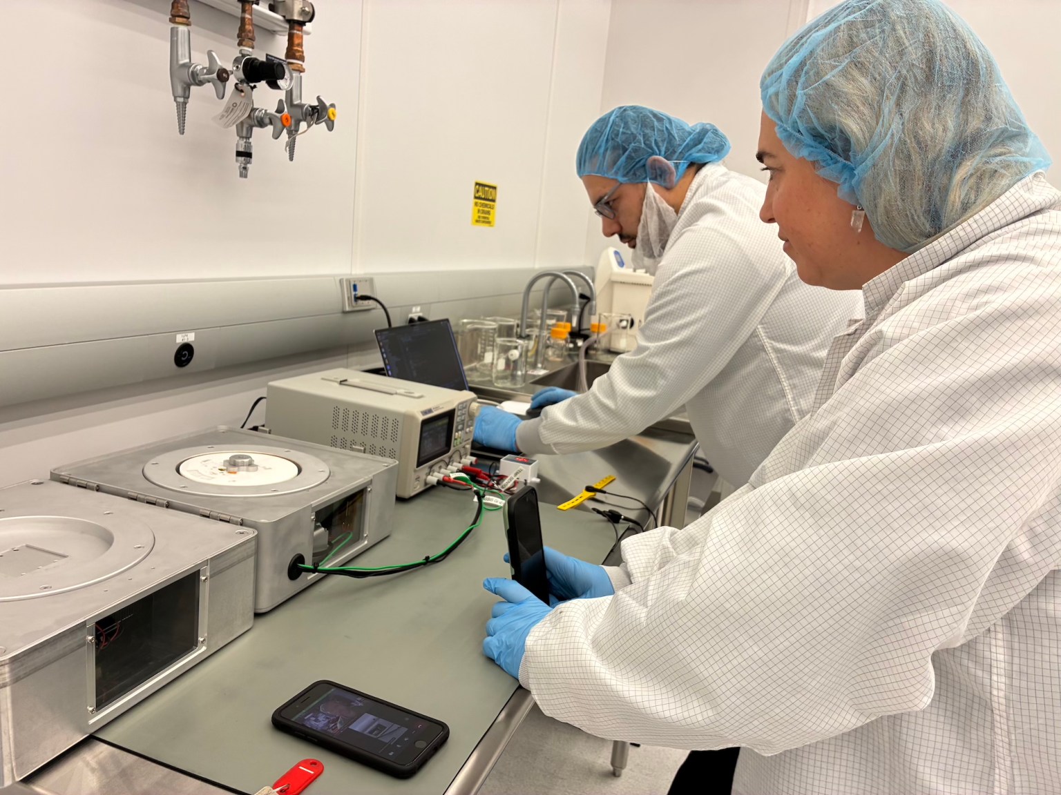 A researcher in a white coats and hair net records an experiment with a cell phone while another researcher types on a laptop.