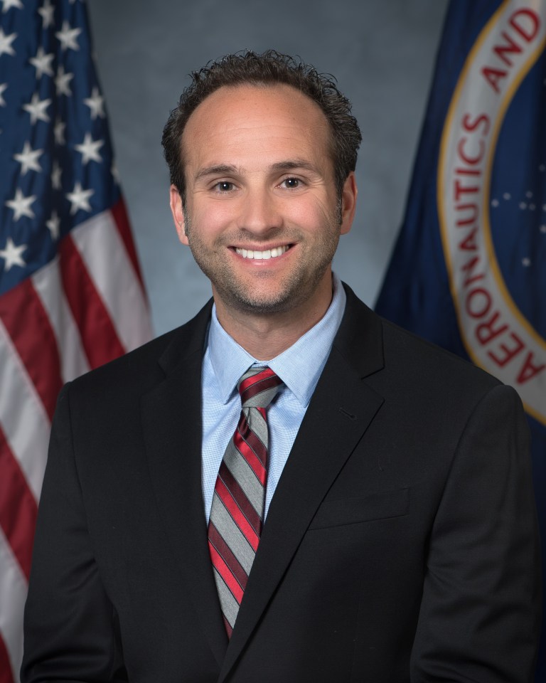 A portrait of Jerry Traster in a suit in front of the American and NASA flags. 