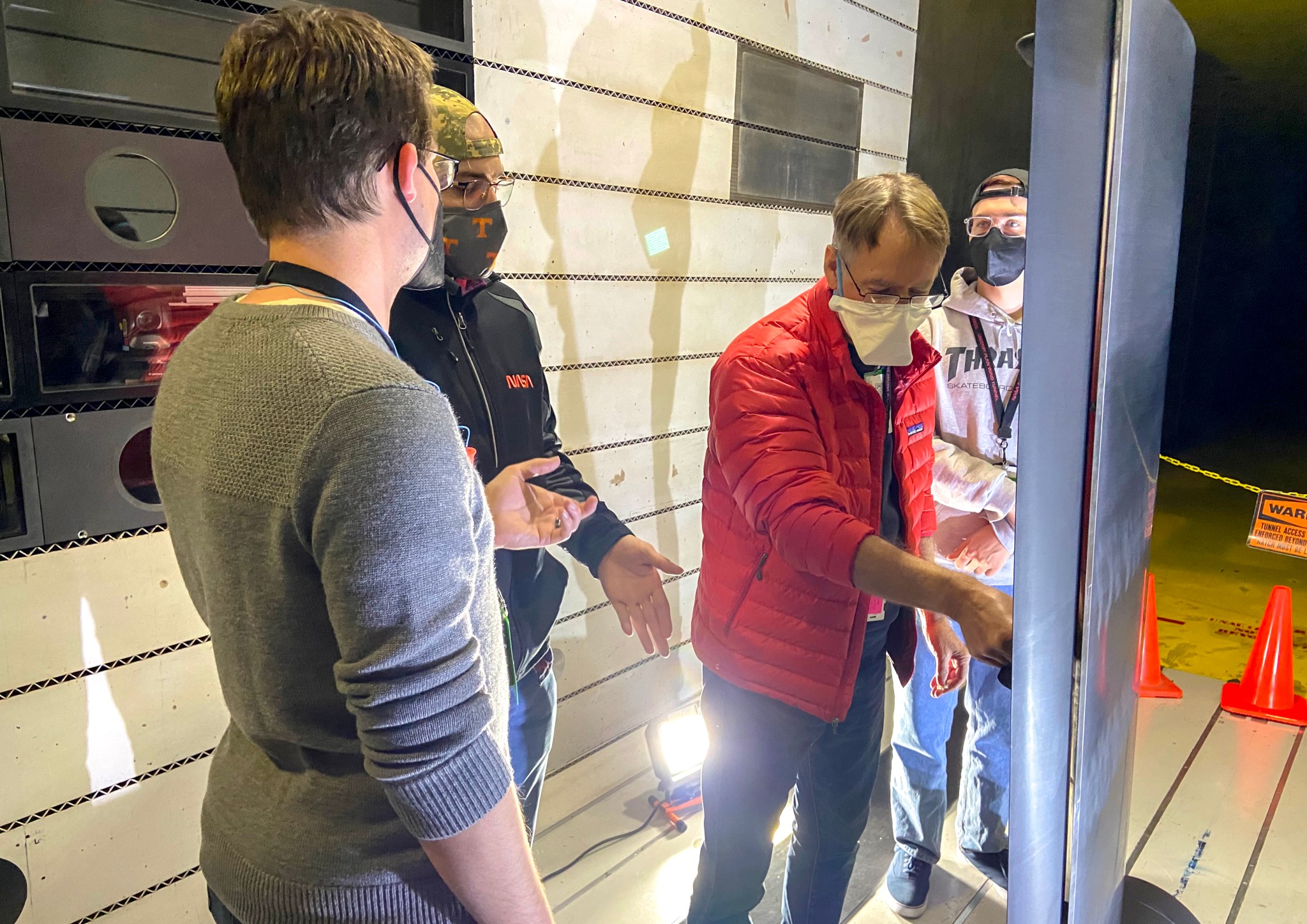 Four men wearing masks stand around a section of an airplane wing mounted vertically inside a NASA wind tunnel as part of a University Leadership Initiative project.