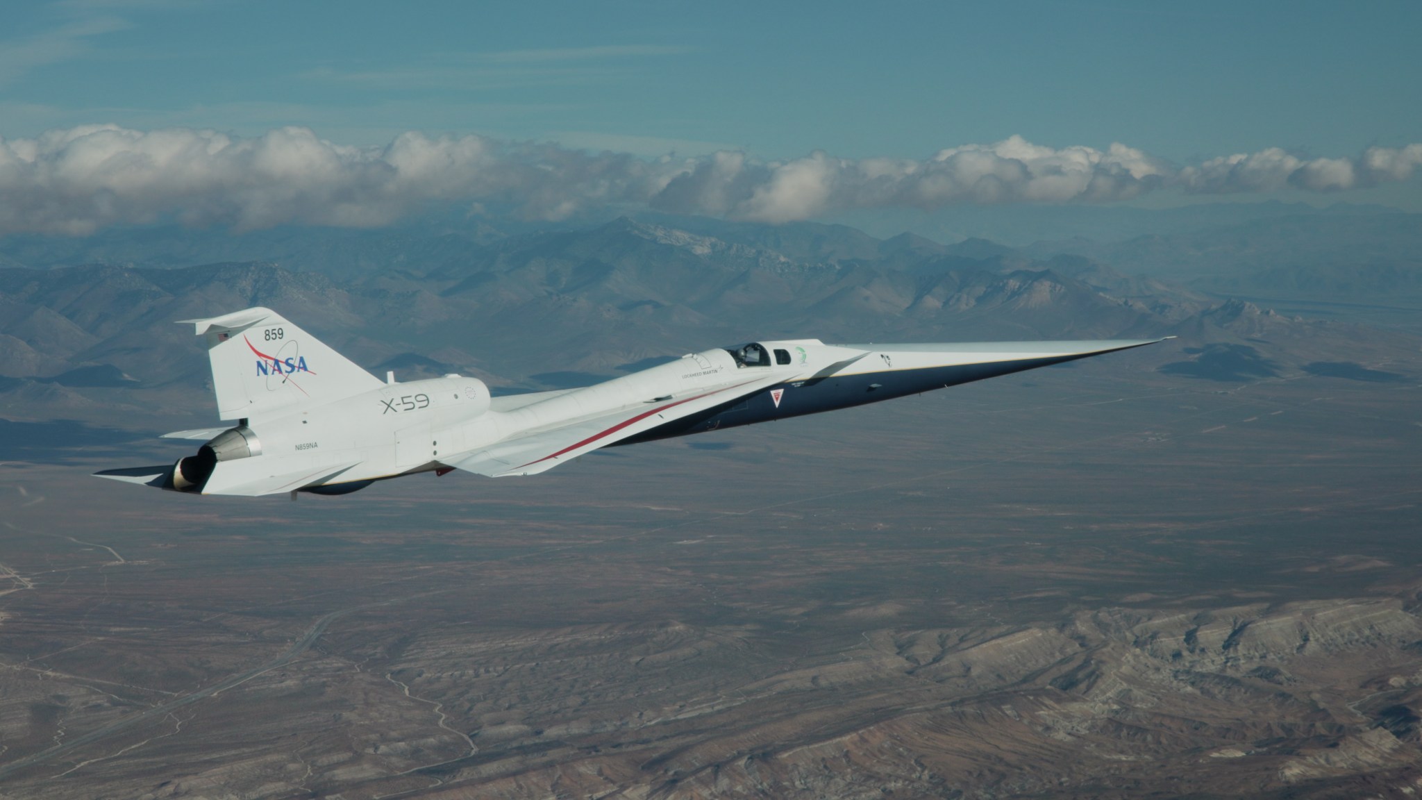 NASA’s X-59 flies above the Mojave Desert on a clear day. The white aircraft has light gray, red, and blue accents, with a NASA logo and the number 859 on its tail. It appears flying level over the desert landscape, with a mountain range visible on the horizon and a trail of clouds above.