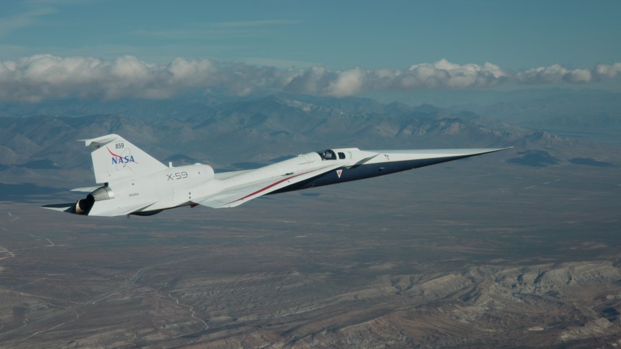 NASA’s X-59 flies above the Mojave Desert on a clear day. The white aircraft has light gray, red, and blue accents, with a NASA logo and the number 859 on its tail. It appears flying level over the desert landscape, with a mountain range visible on the horizon and a trail of clouds above.