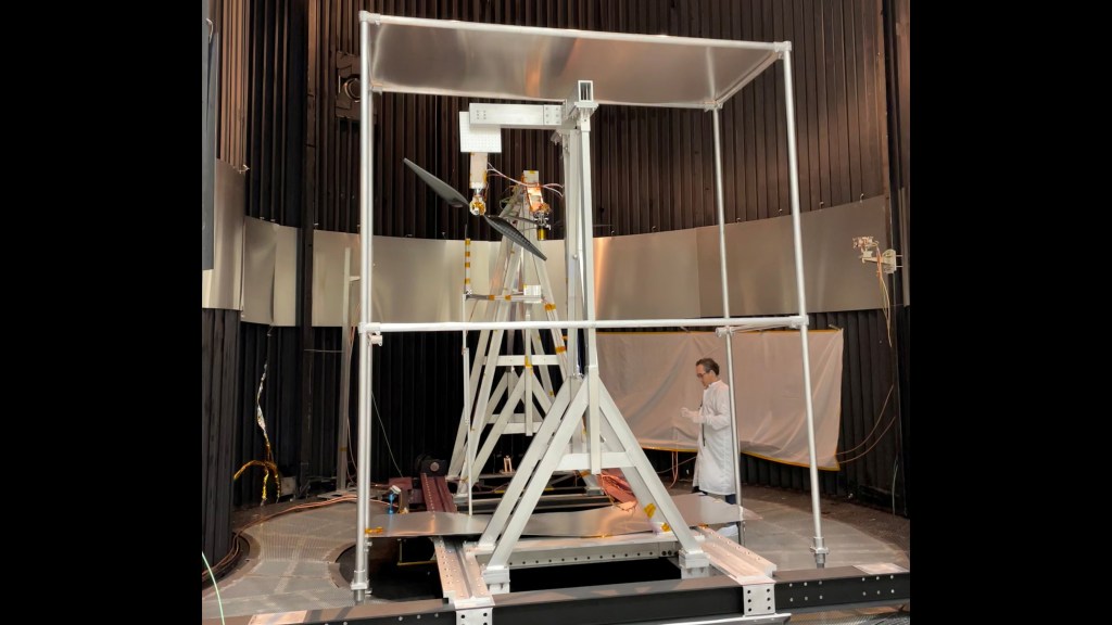 A wide shot inside a dark, cylindrical testing chamber with vertically ribbed walls. In the center, a large silver metal support structure holds a rotor with two long, dark blades. A person in a white lab coat stands to the right of the rig.