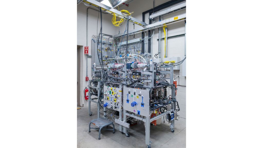 Numerous tubes, wires, and sensors are connected to a large grey system and the wall of a laboratory. A stepstool is visible to the left of the technology, and a fire extinguisher and fire alarm system are seen behind it.