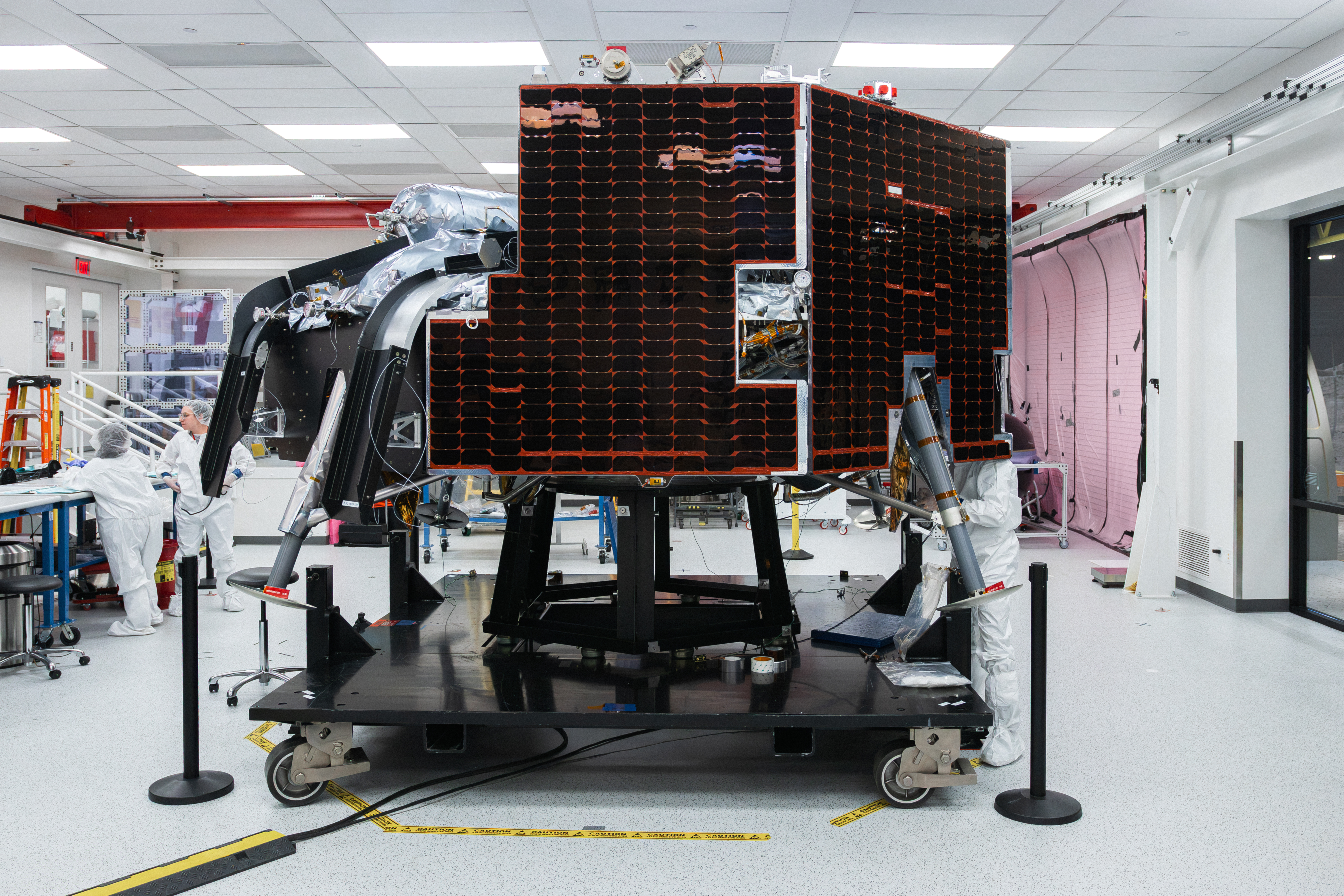 Astrobotic’s Griffin-1 lunar lander sits inside a clean room at the company’s facility. The lander is covered in dark solar panels and mounted on a wheeled test stand. Engineers in white clean-room attire work nearby, with the Astrobotic logo and a large American flag visible in the background.